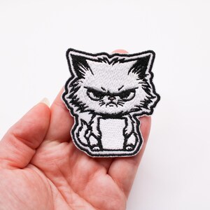 Op de afbeelding: Witte geborduurde strijkpatch met een boze cartoonkat met zwarte contouren.
