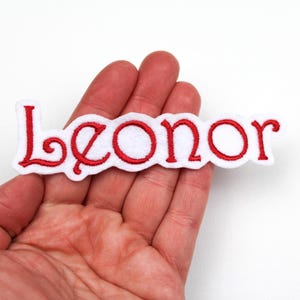 Könnte beinhalten: Ein weißes Filz-Patch mit dem Namen "Leonor" in Rot gestickt, gehalten in einer Hand. Die Schrift ist in einem dekorativen, kursiven Stil mit rotem Rand gehalten.