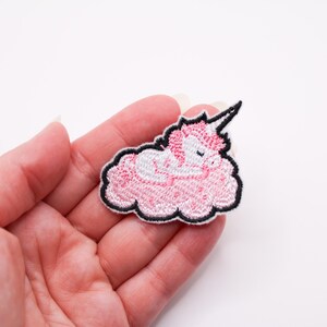 May include: Embroidered patch of a sleeping unicorn on a pink cloud. The unicorn has a white body, pink mane, and a black horn. The cloud and unicorn are outlined in black. The patch is approximately 5 cm long.