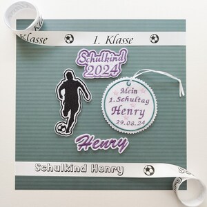 Può includere: Un biglietto verde con un nastro bianco che dice "1. Klasse" e un'icona di pallone da calcio. Ci sono diversi patch ricamati sul biglietto, tra cui un calciatore, un cerchio con il testo "Mein 1. Schultag Henry 29.08.24", e il nome "Henry" in lettere viola. Il testo "Schulkind Henry" è anche stampato sul biglietto.