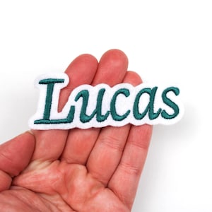 Puede incluir: Parche bordado blanco con el nombre "Lucas" en escritura verde azulado. El parche tiene un borde blanco y está diseñado para ser cosido o planchado sobre tela. El nombre está en una fuente cursiva.