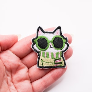 Op de afbeelding: Geborduurde patch met een witte kat met een groene zonnebril en een groen-roze gestreepte sjaal. De kat heeft zwarte ogen en een zwarte omtrek. De patch wordt in een hand gehouden.