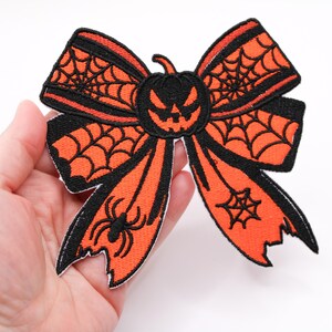 May include: Embroidered Halloween bow patch in orange and black. The bow features a jack-o'-lantern in the center, spiderwebs, and a spider. This decorative patch is perfect for adding a spooky touch to clothing or accessories.