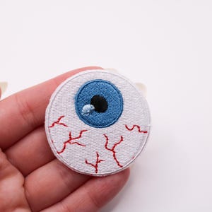 Può includere: Toppa ricamata con un design dettagliato di un occhio. L'occhio ha un'iride blu con una pupilla nera, circondata da una sclera bianca. Le vene rosse sono ricamate intorno all'occhio. Perfetta per abbigliamento o accessori.