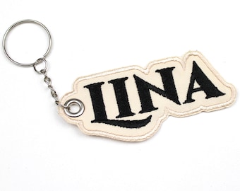 May include: A cream-colored keychain with the name "IINA" embroidered in black. The keychain has a silver metal ring and chain for attaching to keys or bags. The embroidered patch has a decorative border.