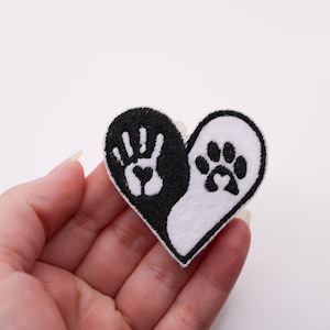 May include: A heart-shaped patch, split into black and white halves. The black side features a white handprint with a heart, while the white side has a black paw print. The patch is outlined in black.