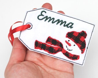 May include: A white tag with the name "Emma" embroidered in green. A snowman with a red and black plaid hat and scarf is embroidered below the name. A red ribbon is attached to the tag.