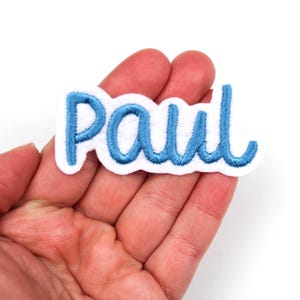 May include: A white felt patch embroidered with the name "Paul" in light blue cursive lettering. The patch has a wavy edge and is held in a hand.