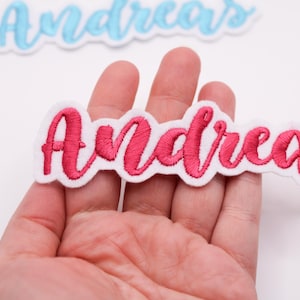 Può includere: Toppa ricamata con il nome "Andrea" in un vivace carattere rosa, bordata di bianco. Una seconda toppa con il nome "Andreas" in azzurro chiaro è visibile sullo sfondo. Le toppe sono adatte per abbigliamento o accessori.