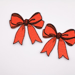 Op de afbeelding: Twee geborduurde strikken in een levendige oranje tint met bruine accenten. De strikken hebben een gestikte textuur en een klassieke strikvorm, tegen een heldere witte achtergrond. Ideaal voor knutselen of versieringen.