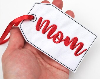 May include: A white tag with the word "Mom" embroidered in red script. The tag has a black stitched border and a red ribbon for hanging. The tag is held in a hand.