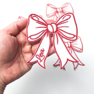 May include: Embroidered white bow patches with red detailing. The patches feature a white bow design with red outlining and the name "Lea" in red script. Several bow patches are visible against a white background.