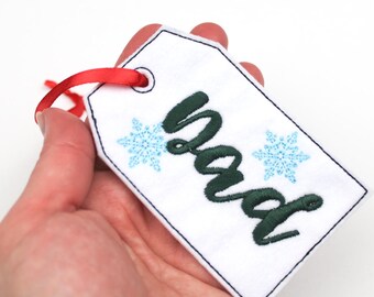 May include: A white gift tag with the word "dad" embroidered in dark green script, flanked by two light blue snowflake designs. The tag has a red ribbon for hanging and a dark blue border.