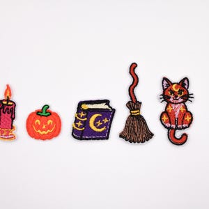 May include: Embroidered Halloween patches including a candle, pumpkin, spell book, broom, and cat. The patches are in orange, purple, and red, set against a white background. Ideal for adding to clothing or accessories.