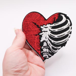 May include: Embroidered patch in the shape of a heart, split vertically. The left side is solid red, while the right side features a black and white rib cage design. The patch is held against a white background.