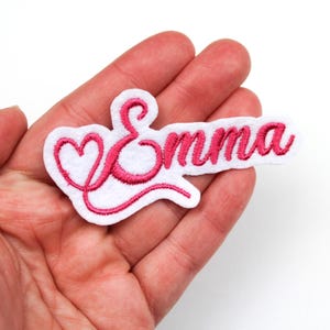 Puede incluir: Un parche de fieltro blanco bordado con el nombre "Emma" en escritura cursiva rosa. La letra "E" está estilizada con forma de corazón. El parche se sostiene en una mano.