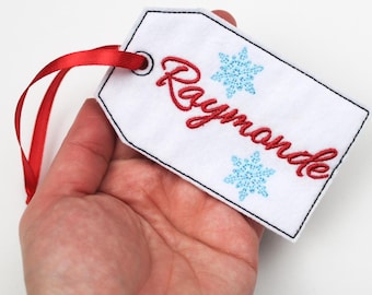 May include: White felt gift tag with the name "Raymonde" embroidered in red script, accented with two light blue snowflake designs. A red ribbon is attached for hanging. The tag has a black stitched border.