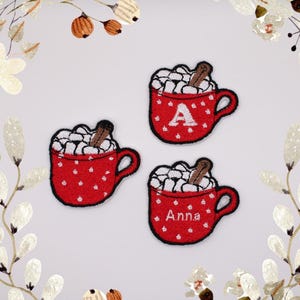 May include: Three red and white embroidered iron-on patches featuring a mug of hot chocolate with marshmallows and a cinnamon stick. The mugs are decorated with white polka dots. One mug has the letter "A" embroidered on it, and another mug has the name "Anna" embroidered on it.