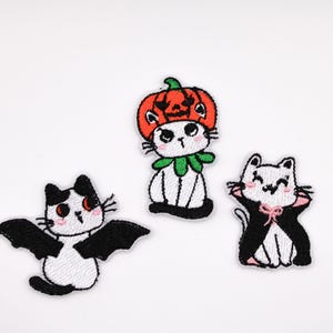 May include: Three embroidered patches featuring cartoon cats in Halloween costumes. One cat wears a pumpkin hat, another has bat wings, and the third wears a cape with a pink bow. The patches are white, black, orange, and green.