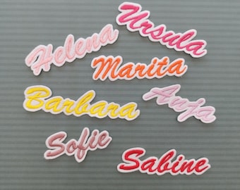 May include: Embroidered name patches in various colors, including pink, orange, yellow, and red. Names include Helena, Ursula, Marita, Barbara, Anja, Sofie, and Sabine, each with a white border. The patches are on a gray striped background.