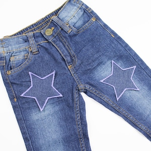 Może przedstawiać: Dżinsy z niebieskiego denimu z dwoma naszywkami w kształcie gwiazd w kolorze fioletowym z przodu.