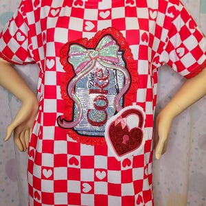 May include: Red and white checkered t-shirt with heart accents. Features a sequined Diet Coke design with a red lace border and a heart-shaped lock patch. The shirt has short sleeves.
