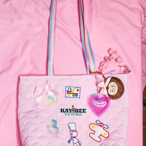 May include: Pink quilted tote bag with rainbow striped handles and embroidered toy-themed patches. Features a bow, a heart charm, and a Kay-Bee Toy Stores logo. Includes a pink teddy bear keychain.