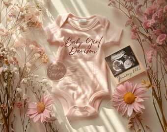 May include: Pink baby onesie with the text "Baby Girl Davison" in maroon cursive. A framed ultrasound image with the date "January 2025" is next to the onesie. Dried flowers are scattered around the onesie and ultrasound image.