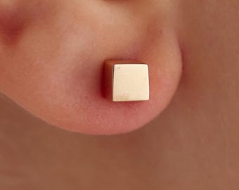 May include: A close-up of a gold square stud earring. The earring is a small, geometric cube shape. The earring is worn on an ear. The earring is a simple, minimalist design.