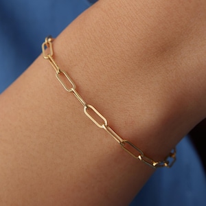 May include: A gold-toned chain bracelet featuring elongated, rectangular links with rounded corners. The bracelet is constructed from a series of uniform, interconnected metal loops that form a continuous, delicate chain design around the wrist.