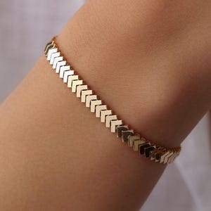 May include: A gold-toned bracelet featuring a continuous chain of interconnected chevron-shaped links. The design consists of repeating V-shaped segments that create a textured, angular pattern along the entire length of the wrist accessory.
