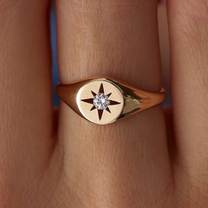 May include: A gold-colored signet ring featuring a starburst design with a central diamond. The ring has a smooth, polished finish and a classic, elegant style. The ring is a piece of jewelry.