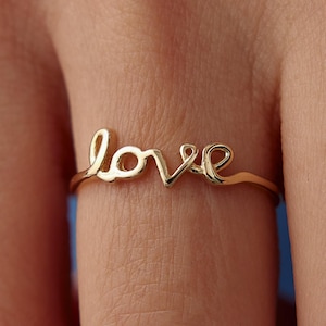 Può includere: Un anello dorato con la parola "love" scritta in corsivo. L'anello è sottile e delicato, con le lettere collegate. L'anello è indossato su un dito, con la pelle visibile sullo sfondo.