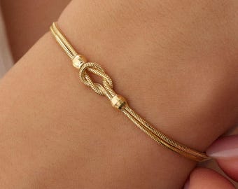 Puede incluir: Una pulsera de tono dorado con un diseño de cadena de doble hebra. El centro de la pulsera presenta un nudo prominente, flanqueado por dos pequeñas cuentas de tono dorado pulido que aseguran la estructura del nudo en su lugar.