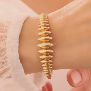 May include: A gold-coloured bracelet featuring a series of elongated, rounded segments. The bangle is worn on a wrist, and the design has a textured surface. The jewellery piece is a bangle style, with a simple clasp.