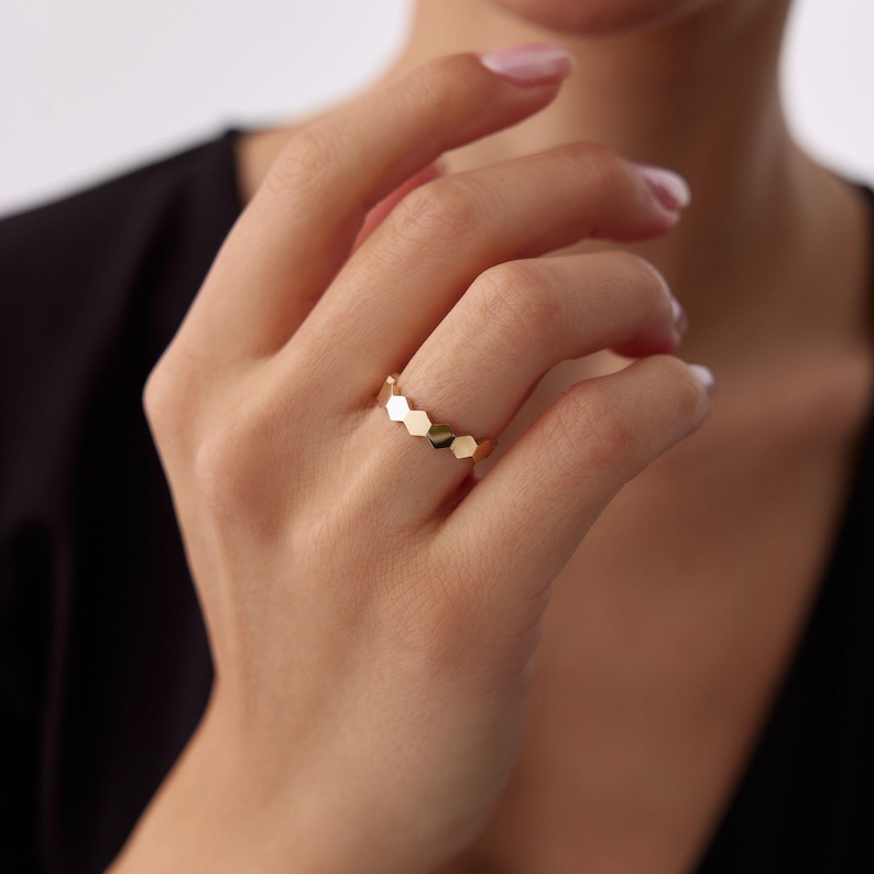 May include: A gold ring featuring a series of connected, geometric hexagonal shapes. The ring is shown on a person's finger, with a black top in the background. The ring's design is modern and minimalist.