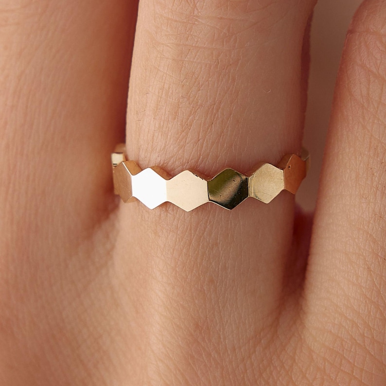 May include: A gold ring with a repeating geometric honeycomb design. The ring is made of a shiny, reflective metal and features a series of connected hexagonal shapes. The ring is shown on a finger, highlighting its delicate and intricate design.