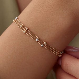May include: A triple-strand bracelet featuring delicate gold-toned ball chains. The design incorporates small, textured, spherical beads in alternating yellow gold and silver tones spaced evenly along the three thin, interconnected metallic chains.