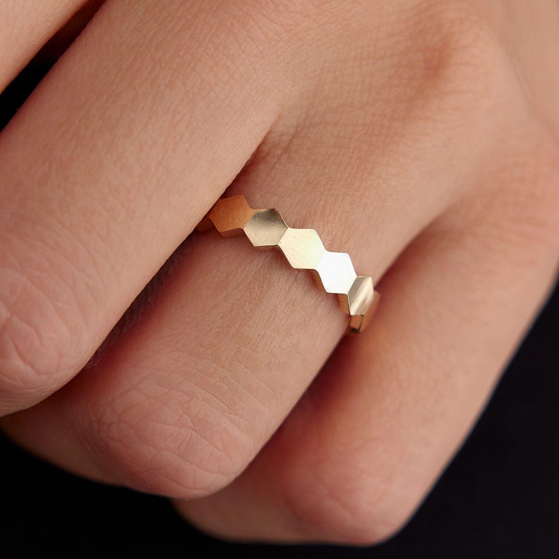 May include: A gold-colored ring with a repeating geometric honeycomb design. The ring is made of a shiny metal and has a series of connected hexagonal shapes. The ring is shown on a finger against a dark background.