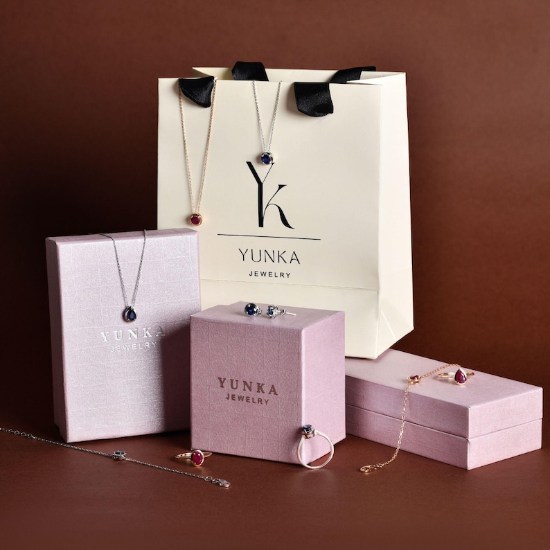 May include: An assortment of jewelry, including necklaces, rings, and bracelets, displayed with gift boxes and a shopping bag. The jewelry features gemstones in red and blue, with the brand name "YUNKA JEWELRY" visible on the boxes and bag.