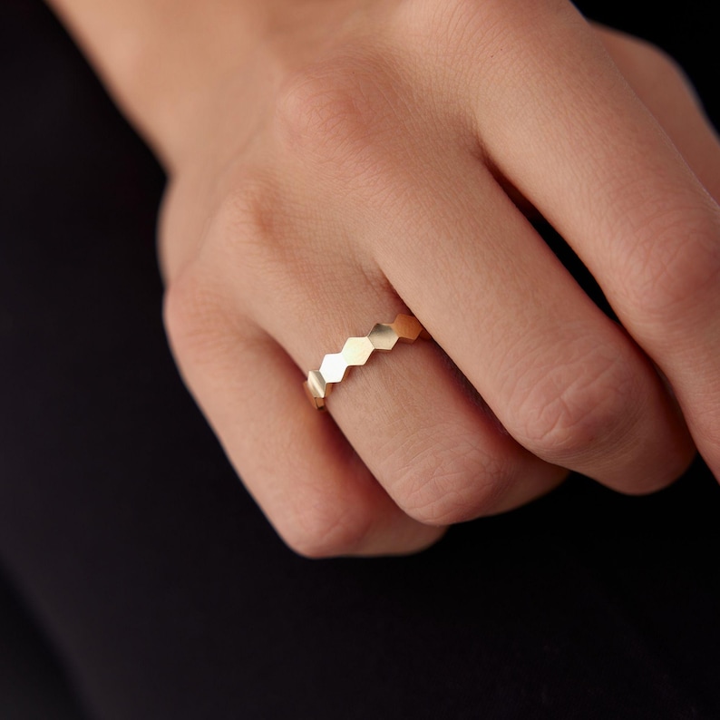 May include: A gold-colored ring with a geometric honeycomb design. The ring features a series of connected hexagonal shapes. The ring is worn on a finger, with the hand and a dark background visible.