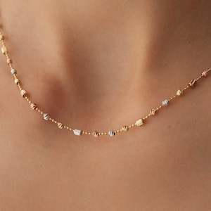 Puede incluir: Un collar de cadena delicado que presenta una serie de pequeñas cuentas texturizadas en tonos alternos de oro amarillo, blanco y rosa. La cadena está compuesta por eslabones finos intercalados con estas esferas y cilindros metálicos pulidos.