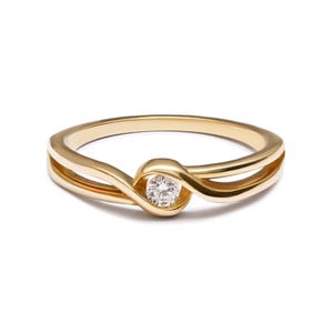 May include: A polished yellow gold ring featuring a bypass-style band that curves to hold a single, round, brilliant-cut clear gemstone at the centre. The band has a smooth, reflective finish and a split design that tapers towards the stone.