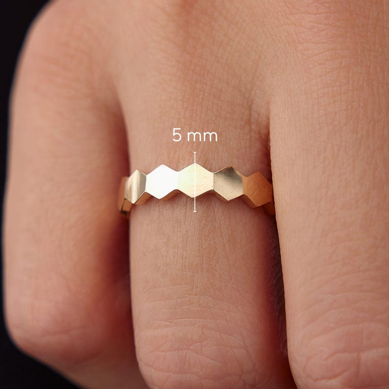 May include: A gold-colored ring with a geometric honeycomb design. The ring is 5 mm wide and features a series of interconnected hexagonal shapes. The ring is shown on a person's finger.