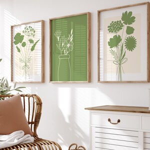 Könnte beinhalten: Ein weißer Korbstuhl mit einem braunen Kissen und einer weißen Wolldecke. Der Stuhl steht vor einer weißen Wand mit drei gerahmten Drucken von grünem Laub. Rechts vom Stuhl steht ein weißer Schrank mit zwei Schubladen und einer Holzplatte.