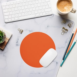 May include: A rectangular desk mat featuring a white marble pattern with grey veining. Centred on the mat is a large, solid orange circle. The mat is surrounded by a white computer keyboard, a white computer mouse, a coffee mug, and pencils.