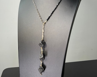 Pode incluir: Um colar preto e dourado exposto em um manequim preto. O colar apresenta uma delicada corrente dourada, uma fileira de pequenas contas pretas e uma queda vertical com três pingentes pretos esculpidos em forma de cubo. Os pingentes são conectados por correntes douradas.