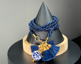 Pode incluir: Uma pulseira de corda azul marinho com detalhes dourados e borlas azul marinho. A pulseira apresenta uma conta decorativa de cerâmica azul e branca. A pulseira é exibida em um suporte preto em forma de cone.