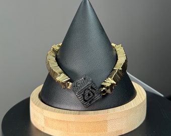 Pode incluir: Uma pulseira dourada com um design geométrico, com uma peça central quadrada preta com símbolos esculpidos. A pulseira é exibida em um suporte cônico preto com base de madeira, em um fundo cinza.