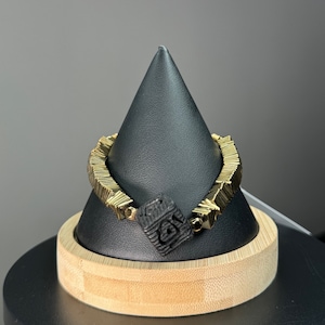 Puede incluir: Una pulsera dorada con un diseño geométrico, con una pieza central cuadrada negra con símbolos tallados. La pulsera se exhibe sobre un soporte cónico negro con base de madera, sobre un fondo gris.